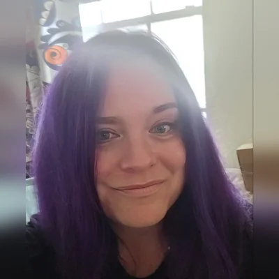 Nicolette, with long purple hair, smiling by a sunlit window
